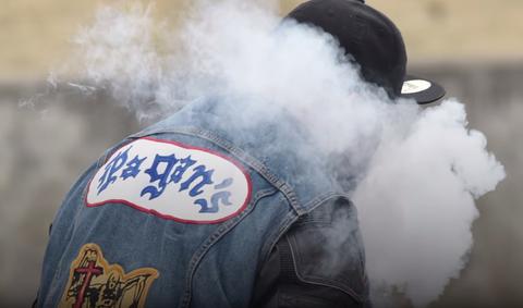 Pagan's MC biker smoking with back turned, wearing a black baseball cap.